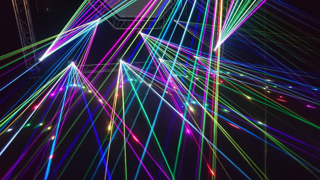 Multiple intersecting beams of colorful laser lights in green, blue, pink, and purple projected in various directions against a dark background.