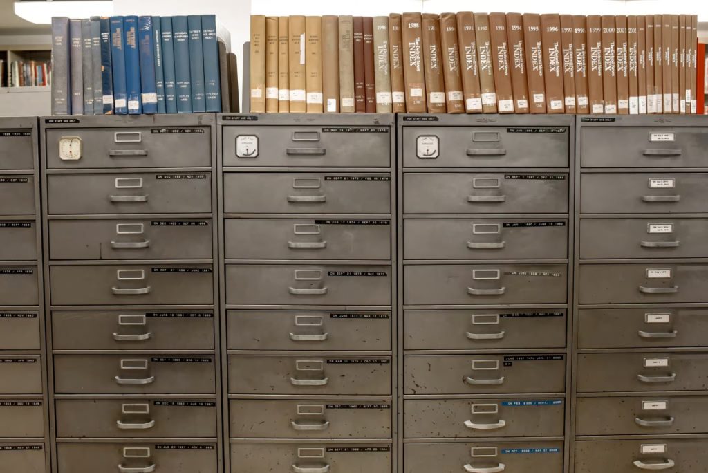 Rows of metal filing cabinets with labeled drawers, topped with blue and brown hardcover library books, in an indoor setting.
