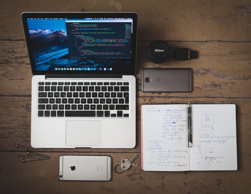 A laptop with code on screen, a camera lens, two smartphones, keys, a pen, and an open notebook with handwritten notes arranged on a wooden table.