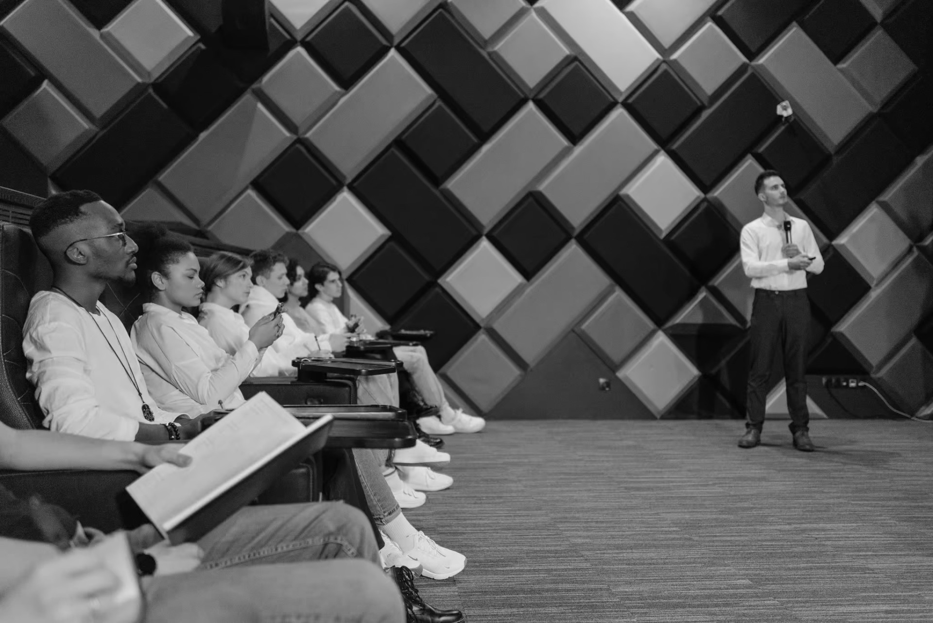 A group of people seated in an auditorium listen to a person standing and speaking in front of a wall with geometric acoustic panels.