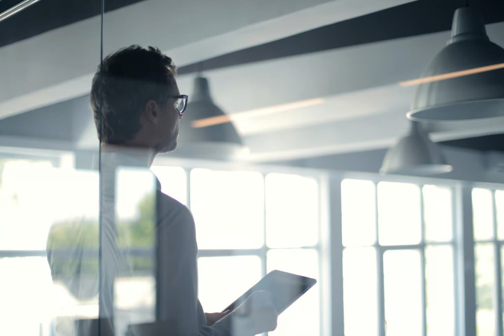 A person holding a tablet stands in a modern office with large windows, looking out.