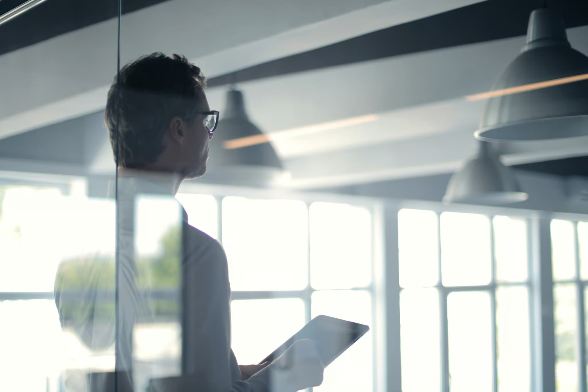 A person holding a tablet stands in a modern office with large windows, looking out.