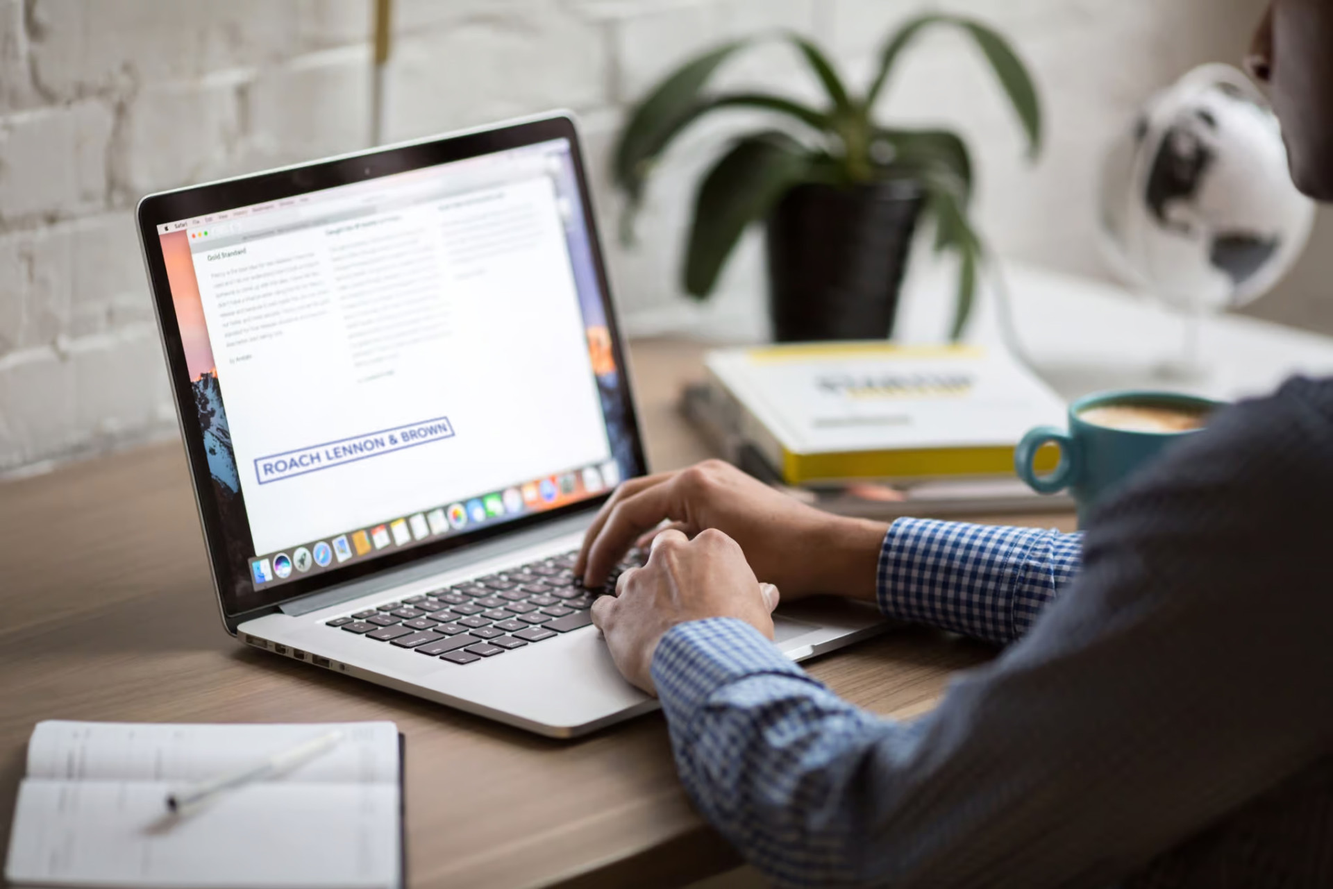 Person typing on a laptop at a wooden desk with an open notebook, a stack of books, and a potted plant in the background.