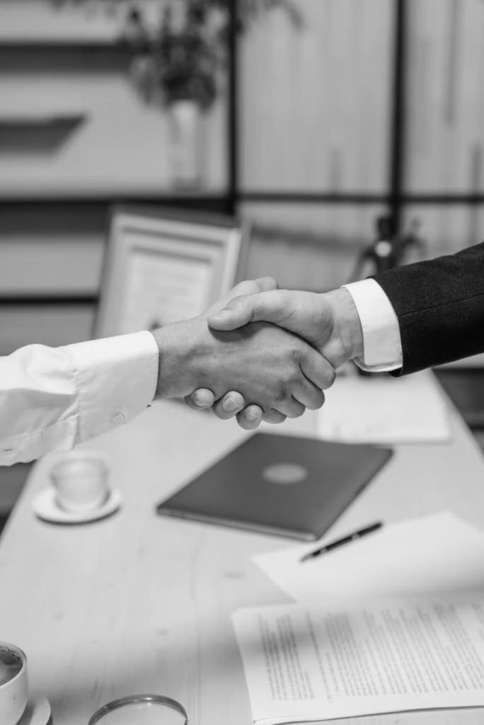 Two people in business attire shaking hands over a desk with documents, a laptop, and a coffee cup, suggesting a professional agreement or meeting.