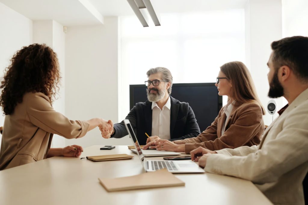 Four people in business attire sit at a meeting table, with two of them shaking hands, suggesting agreement or introduction, while the others observe.