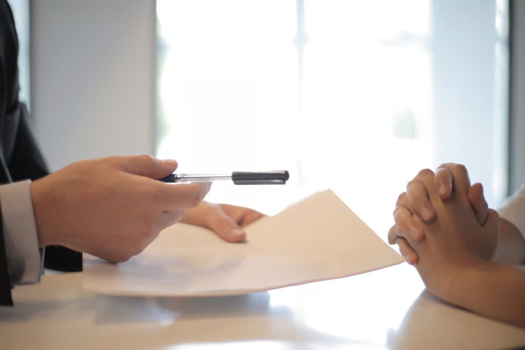 Two people sit at a table; one person hands a pen and paper to the other, whose hands are clasped together.