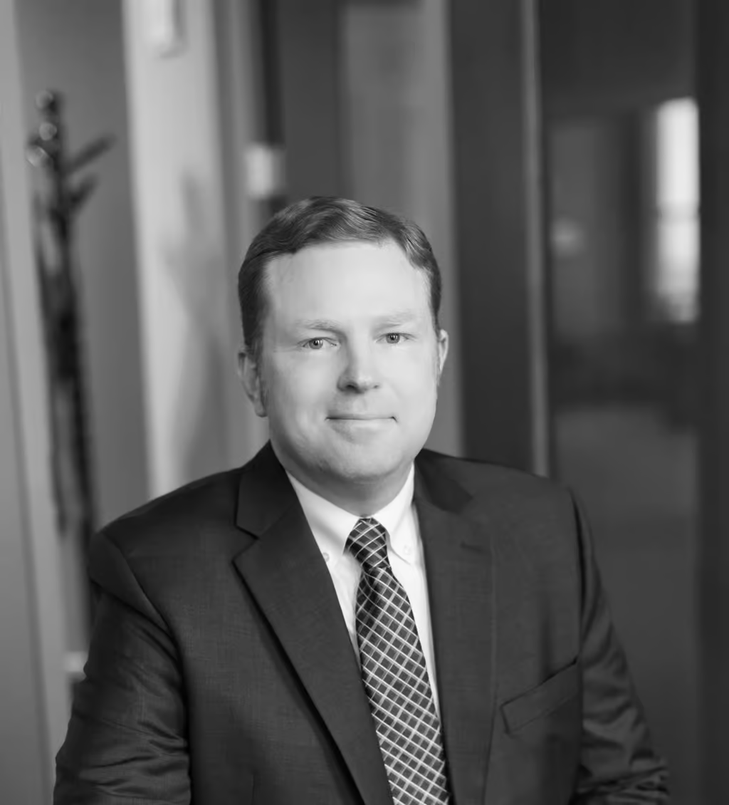 A man in a suit and tie sits in an office environment, looking at the camera with a neutral expression.