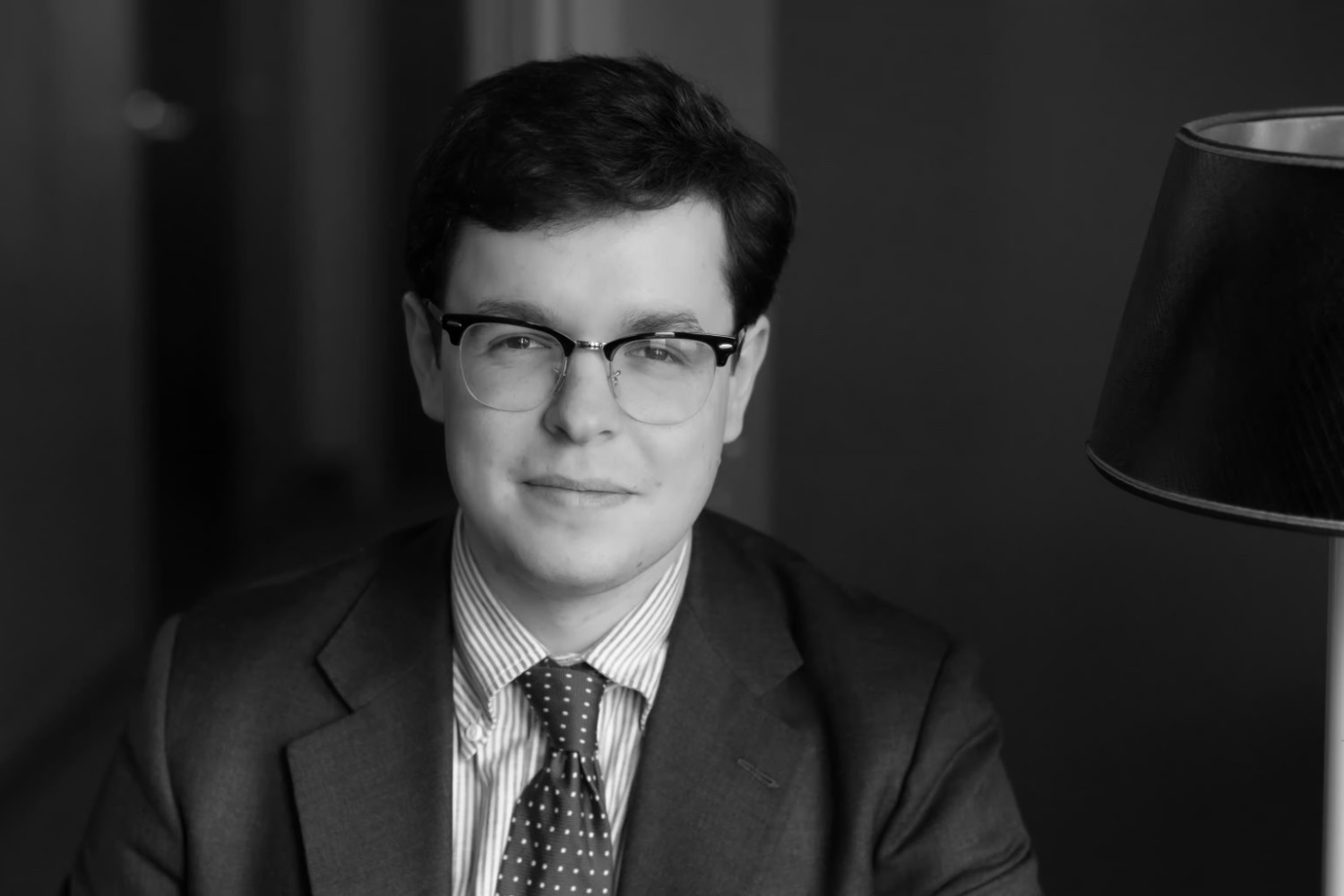 A man wearing glasses, a suit, and a striped shirt with a polka dot tie sits indoors next to a lamp, facing the camera with a neutral expression.