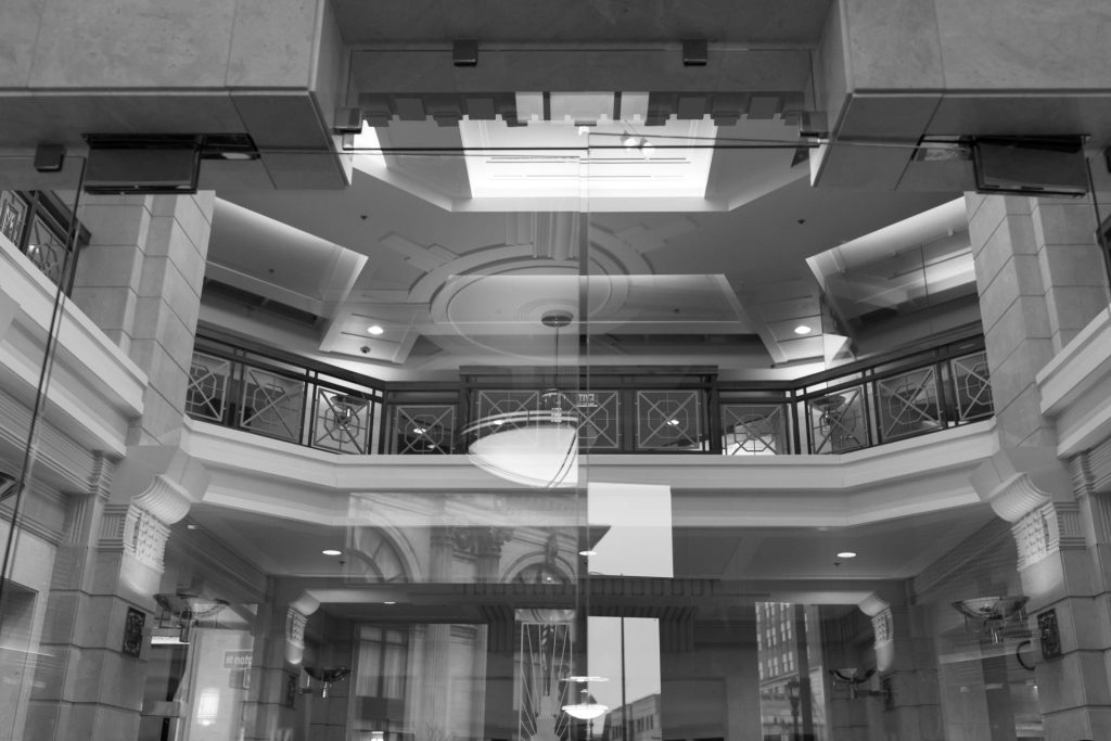 Black and white photo of a building interior featuring a glass wall, decorative ceiling, columns, railings, and reflections of windows and city structures.