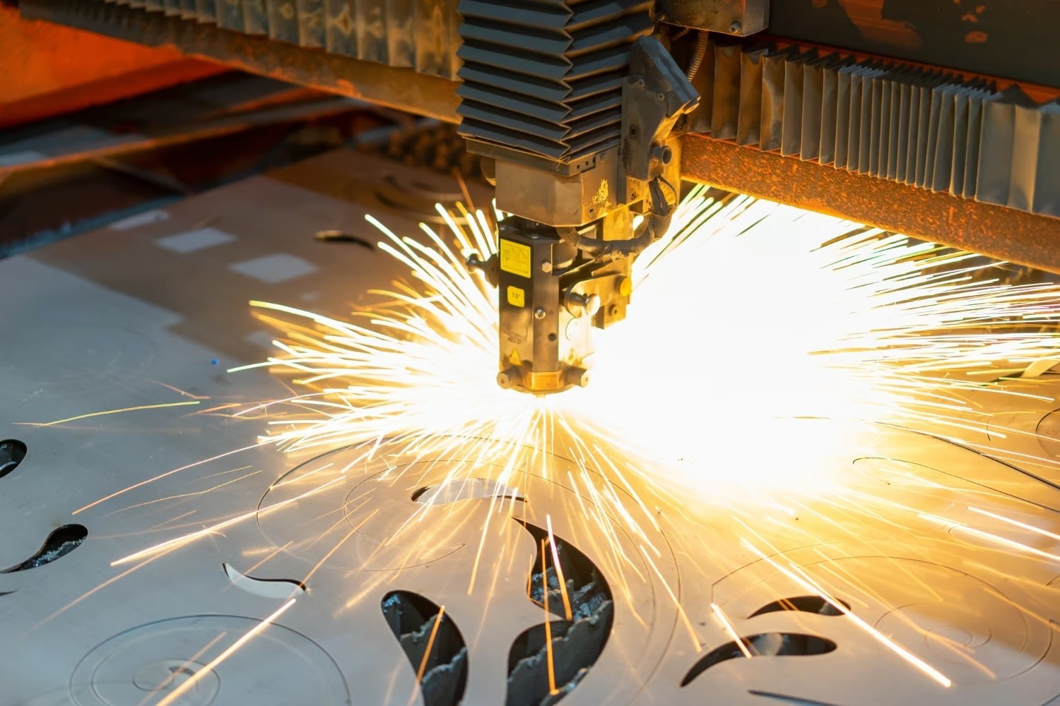 A laser cutting machine emits bright sparks while cutting intricate shapes into a sheet of metal in an industrial setting.