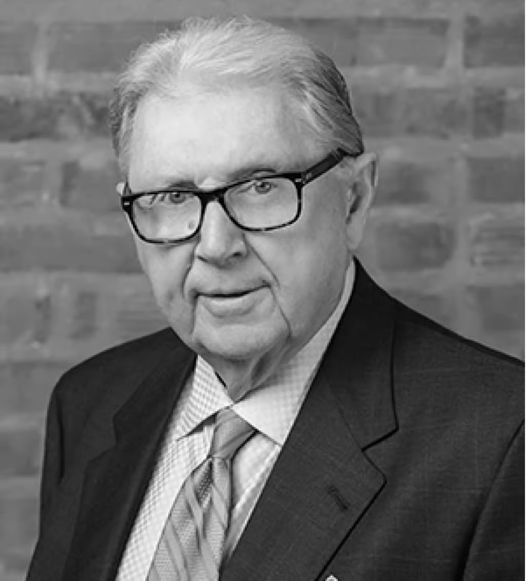 Elderly man with gray hair and glasses wearing a suit and tie, posed in front of a brick wall.