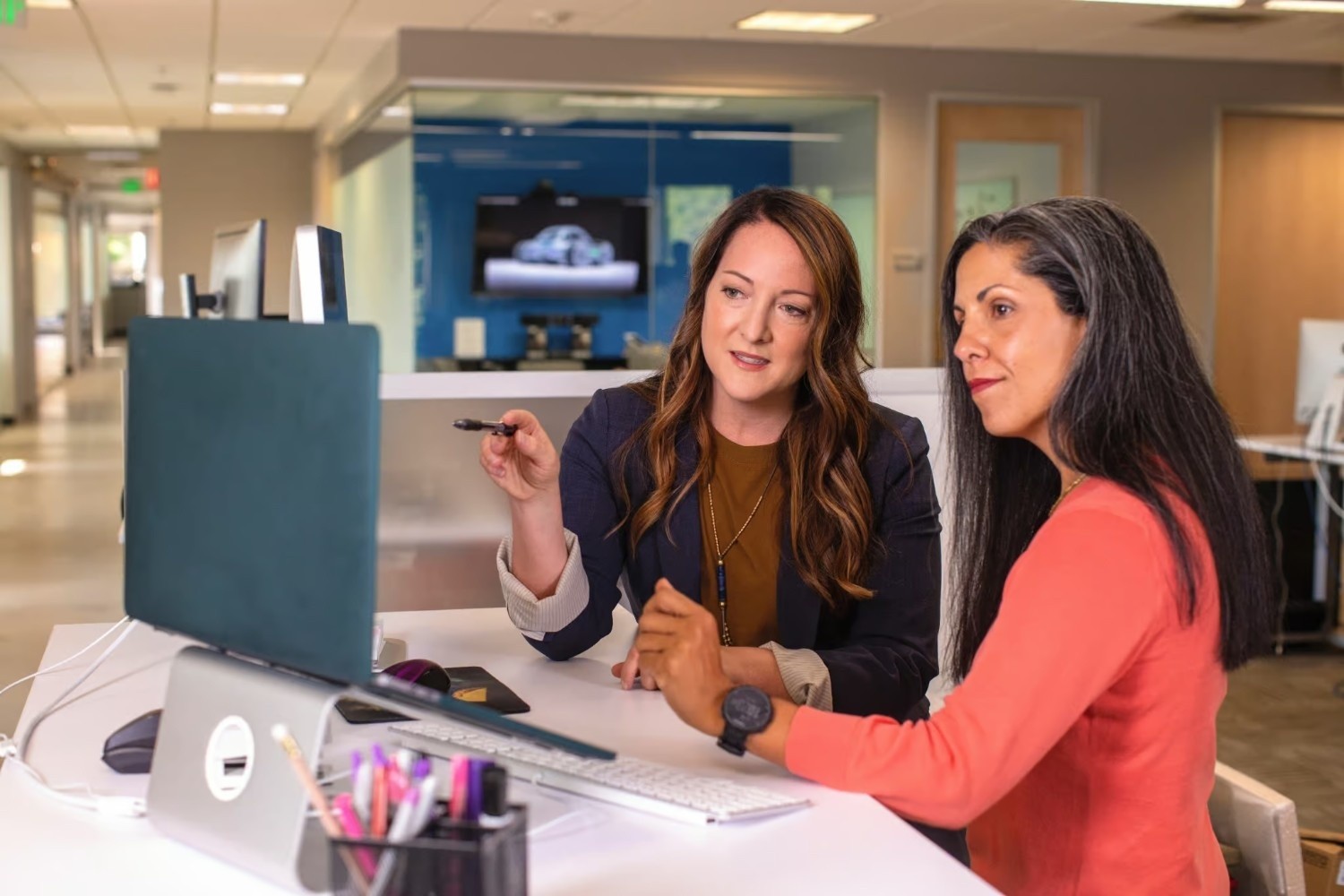 Two women sit at a desk in an office, looking at a computer monitor. One woman is pointing at the screen with a pen while the other listens attentively.