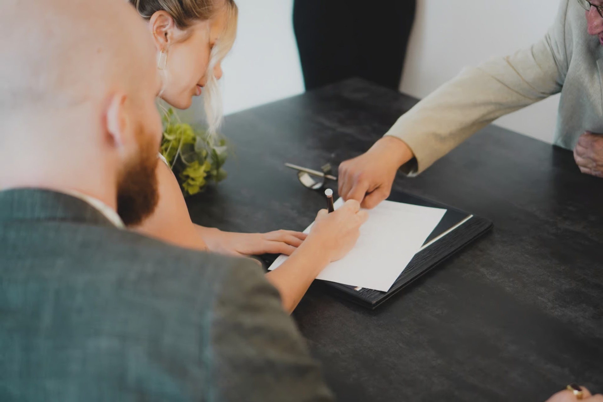 A woman signs a document on a clipboard at a table, while three other people are present, one pointing at the paper.