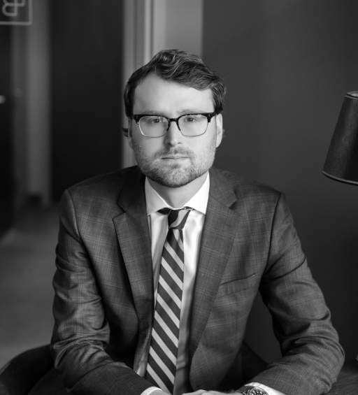 A man in a suit and glasses sits indoors, looking at the camera with a neutral expression. A lamp is visible to his right. The image is in black and white.