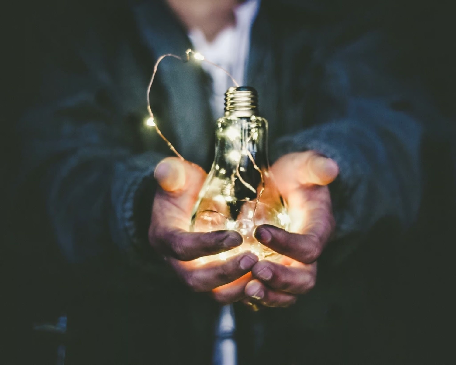 A person holds a clear lightbulb filled with small glowing string lights in both hands, against a dark background.