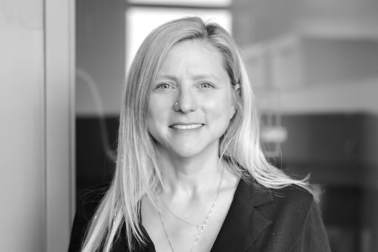 A woman with long blonde hair wearing a dark blazer smiles at the camera in an indoor office setting.