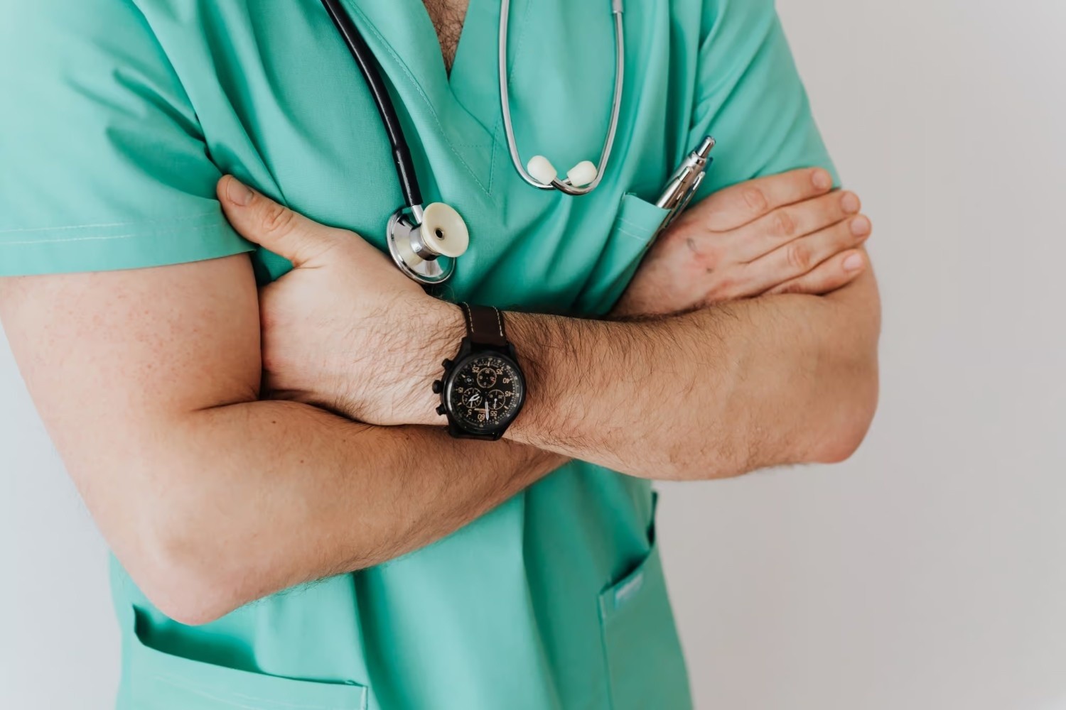 Person in green medical scrubs with a stethoscope around their neck, arms crossed, and a pen in a pocket. Face is not visible.