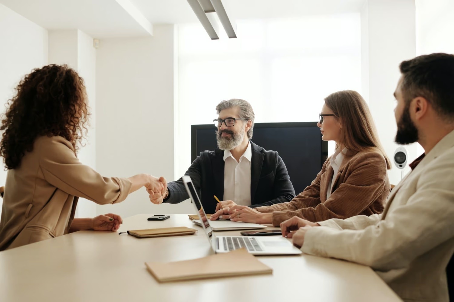 Three people sit at a conference table with laptops and documents while a woman shakes hands with a man across the table in a bright office setting.