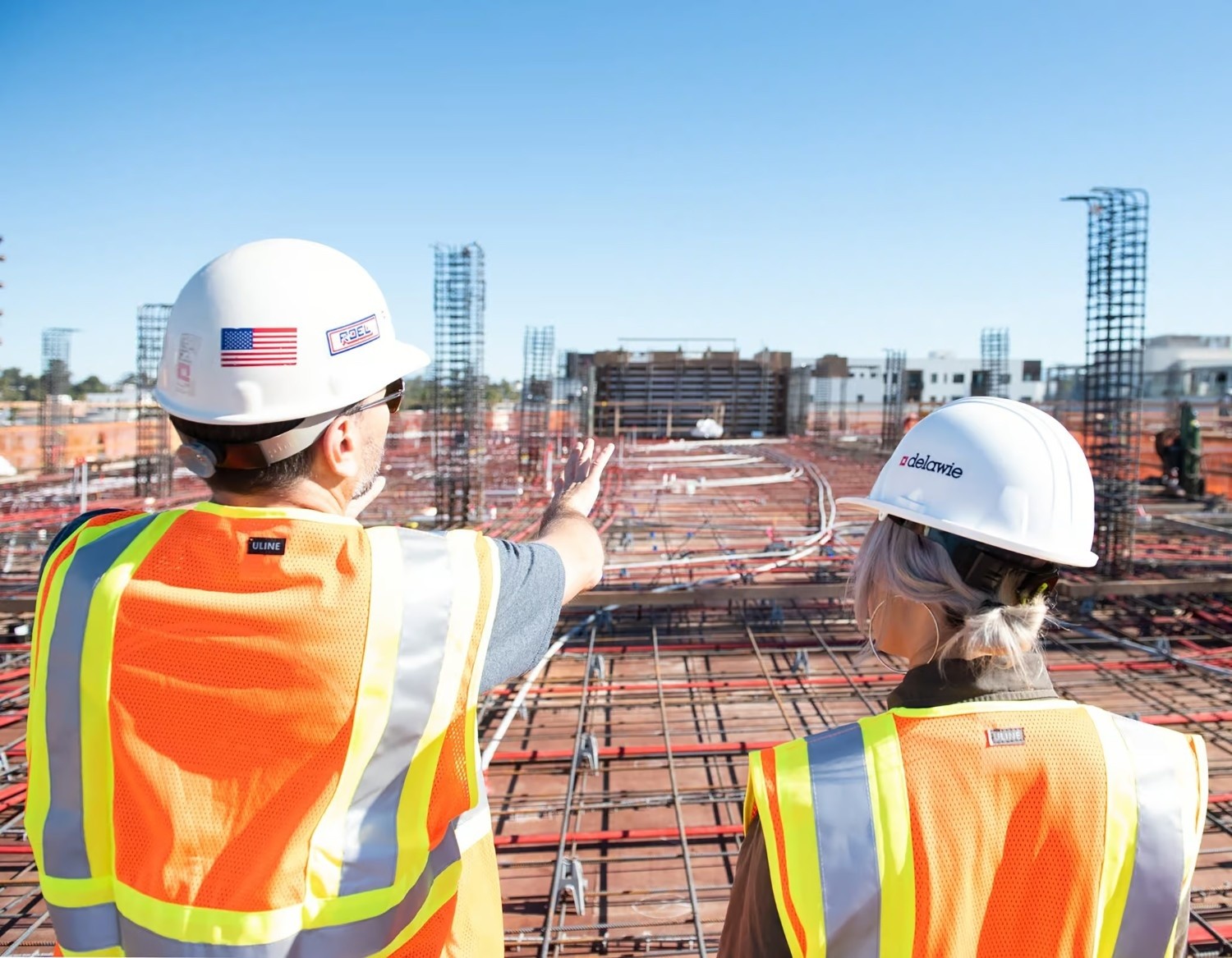 Two construction workers in safety vests and hard hats stand on a building site, with one pointing toward the steel framework and rebar of an unfinished structure.