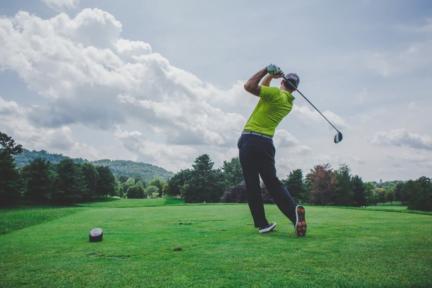 A golfer in a yellow shirt and black pants swings a golf club on a green fairway under a partly cloudy sky. Trees and hills are visible in the background.
