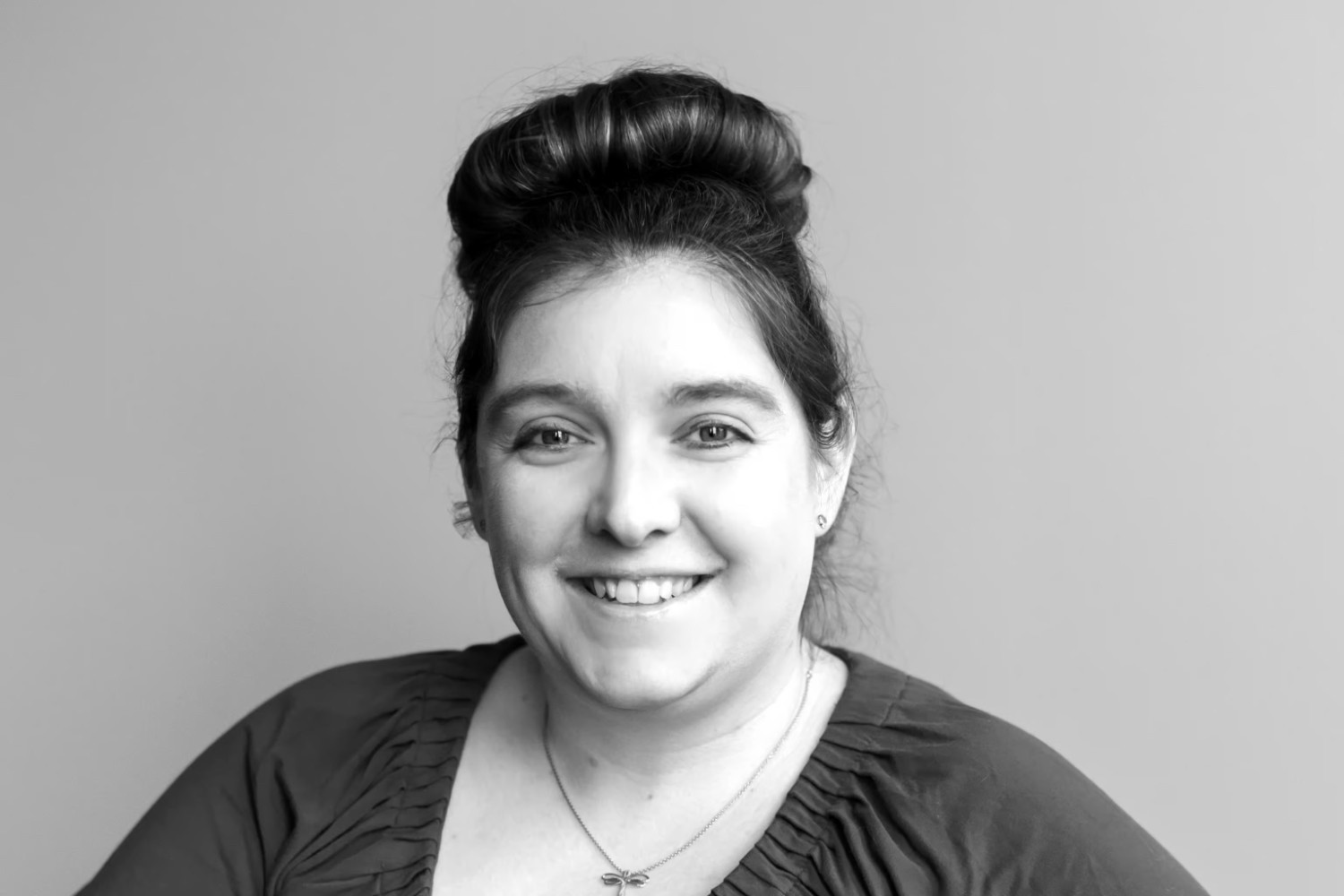 A woman with dark hair in an updo smiling at the camera, wearing a dark top and small earrings, against a plain background.