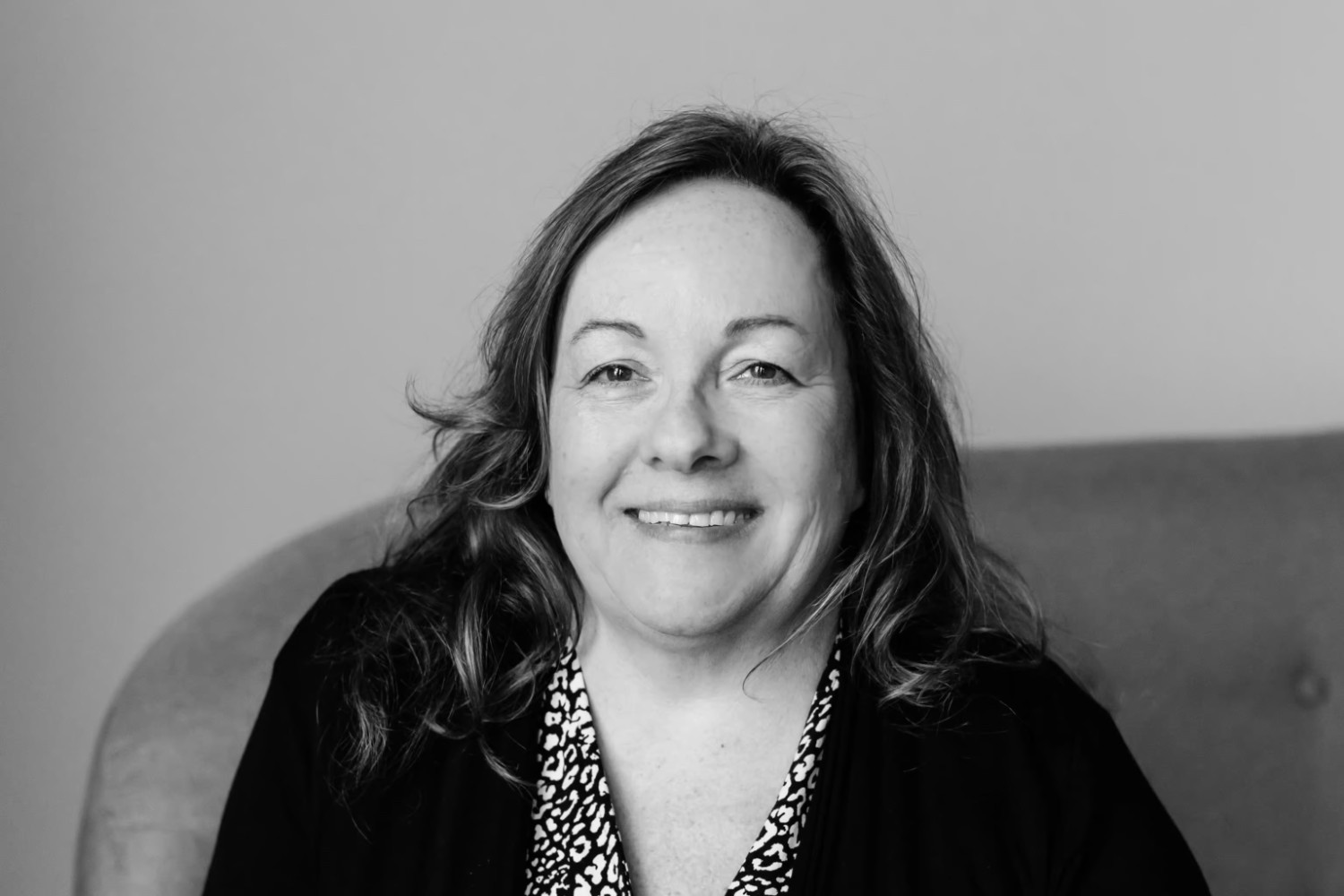 A woman with wavy hair sits on a cushioned chair, smiling at the camera in a black and white photo.