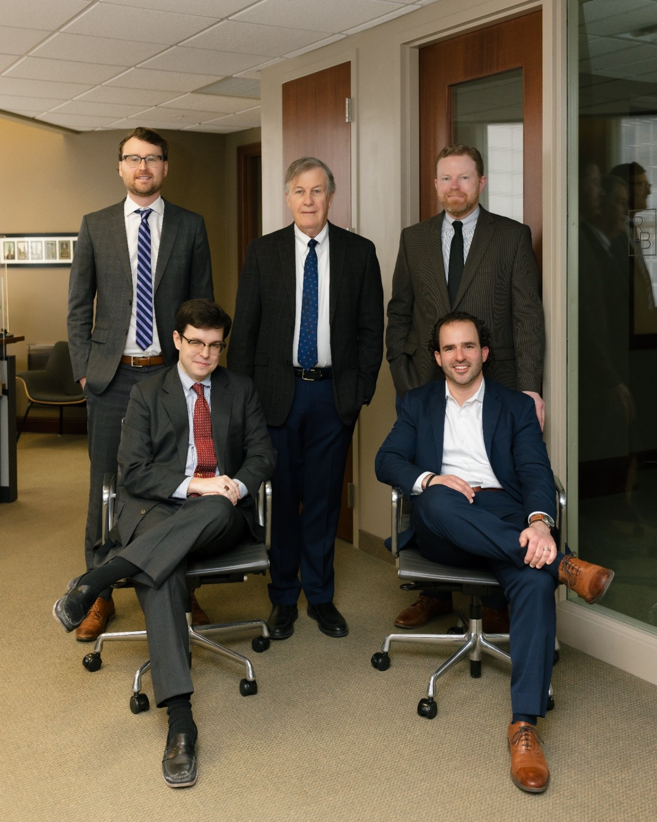Five men in business attire pose in an office hallway; three are standing and two are seated on office chairs, looking at the camera.