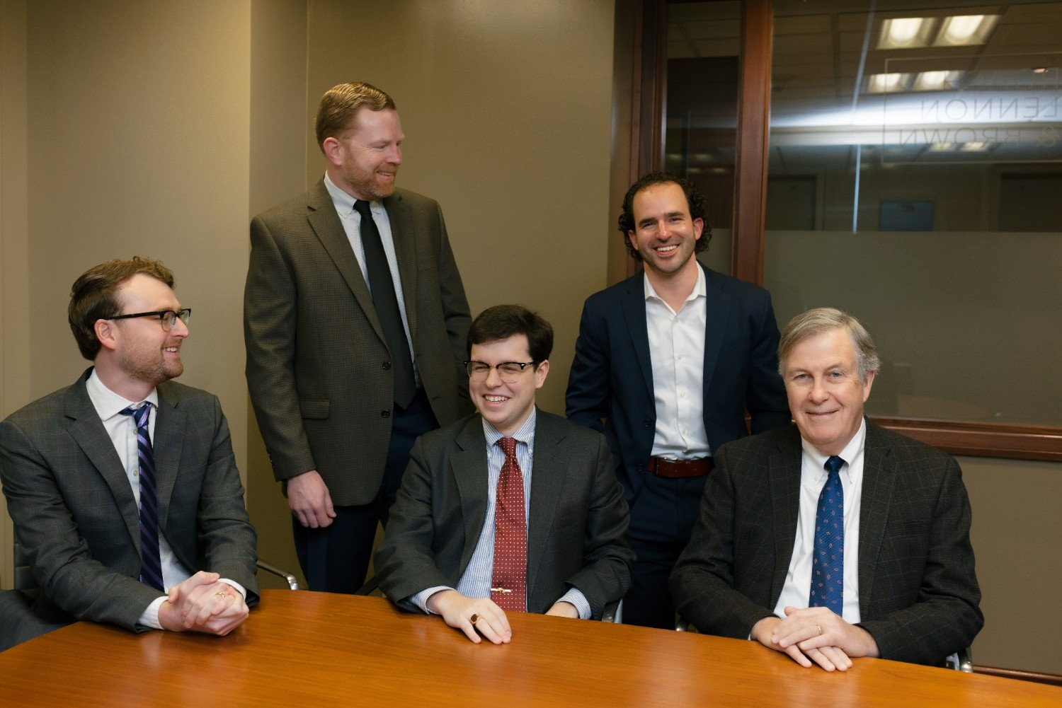 Five men in business attire are gathered around a conference table, with three seated and two standing, all smiling or looking engaged in an office setting.