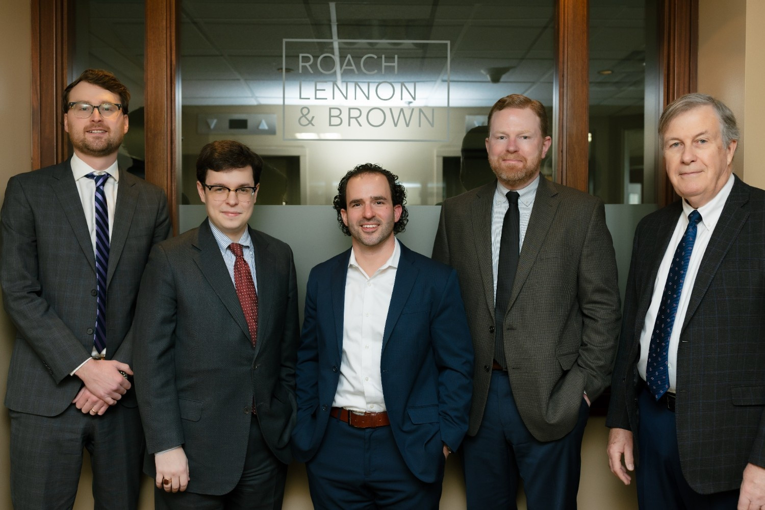 Five men in business attire stand in front of a glass door with the sign "Roach Lennon & Brown" visible in an office setting.