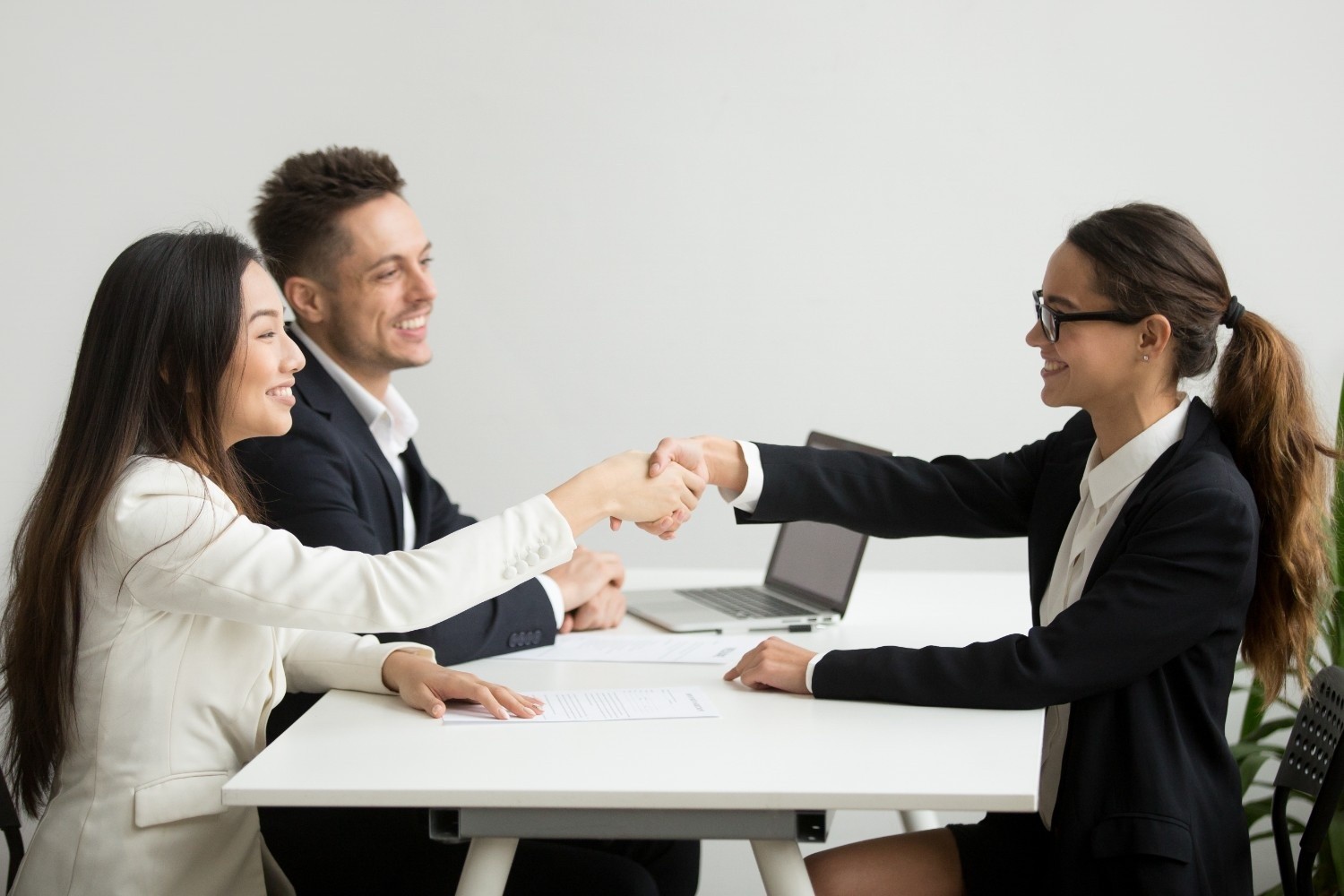Three people in business attire sit at a table; two women shake hands while a man looks on, all appearing engaged in a professional meeting.
