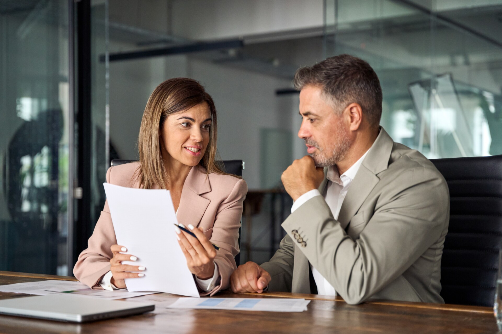 Two professionally dressed people sit at a desk in an office, discussing documents. One person holds papers and a pen, while the other listens attentively.